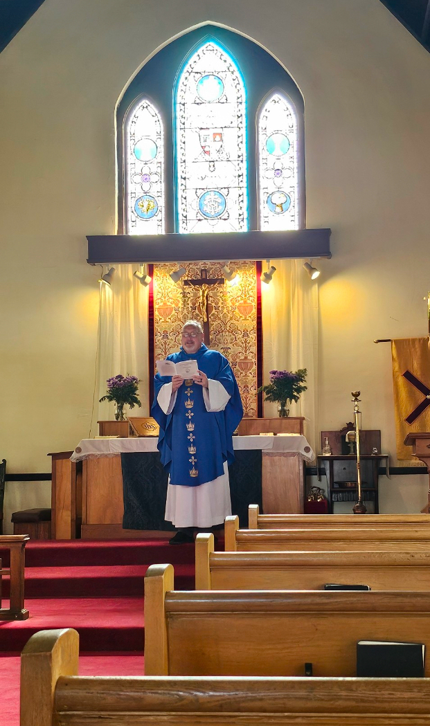 Auto-generated description: A priest in blue vestments stands at an altar in a church with a stained glass window above him.